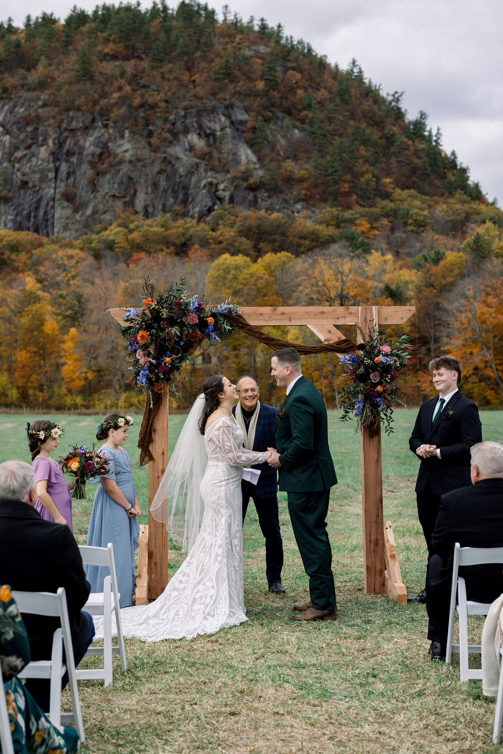 Bride and groom exchanging vows under a floral arbor with White Mountains cliffs and fall foliage at Alpine Garden